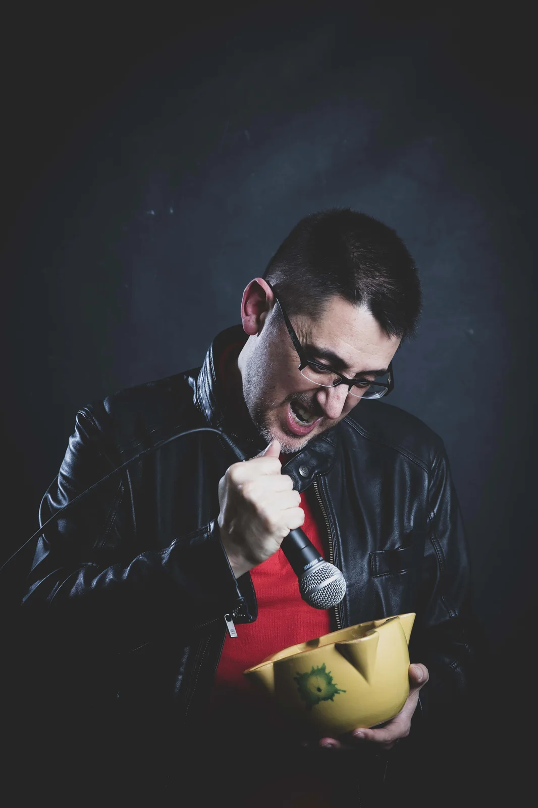 Marcos Félix con chaqueta de cuero, polo rojo y usando el micrófono como mazo de mortero